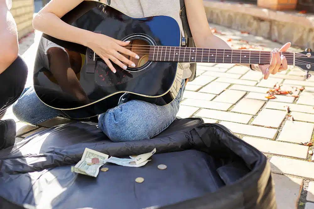 Close-up of girl’s hand playing acoustic guitar and case with cash on sidewalk De jeunes musiciens dans la rue. Gros plan sur la main d’une fille jouant de la guitare acoustique avec une valise contenant de l’argent liquide posée devant elle sur le trottoir.