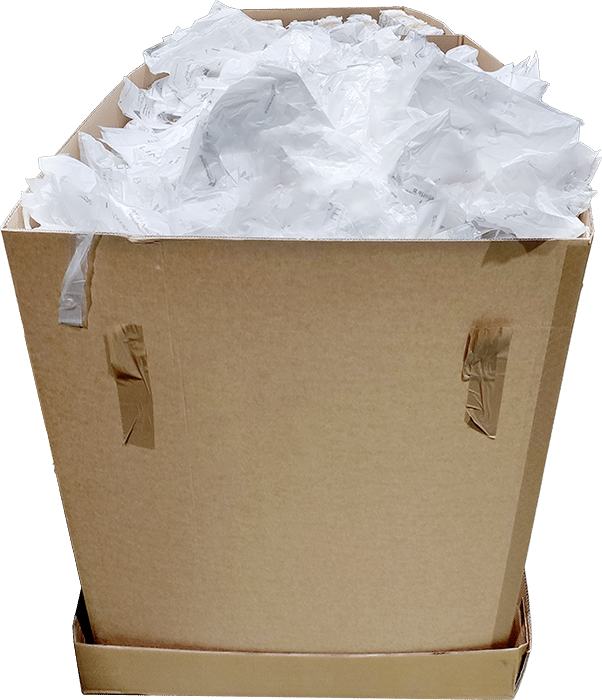Un carton débordant de pochettes plastiques blanches en vrac et froissées, illustrant l’état initial des déchets plastiques collectés avant le compactage.