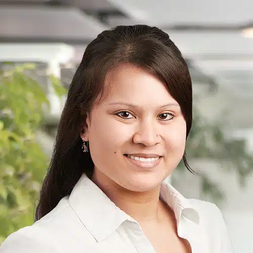 Gros plan d’Alexandra Böhmer, Département Achats chez Debatin. Une femme souriante avec de longs cheveux foncés et un piercing au nez. Elle porte un chemisier blanc, avec des plantes vertes et des structures claires floues en arrière-plan.