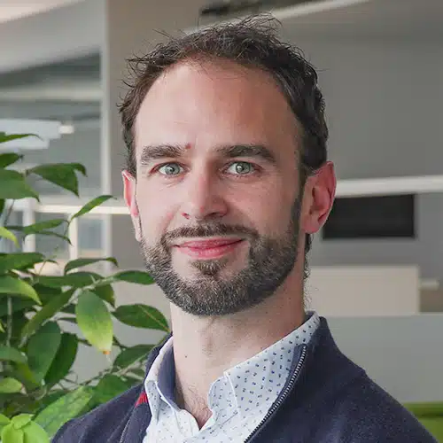 Portrait de Christophe Pille, un homme aux cheveux foncés bouclés et à la barbe courte. Il porte une chemise bleu clair à motifs et un pull sombre. Christophe Pille travaille en Support commercial chez DEBATIN.