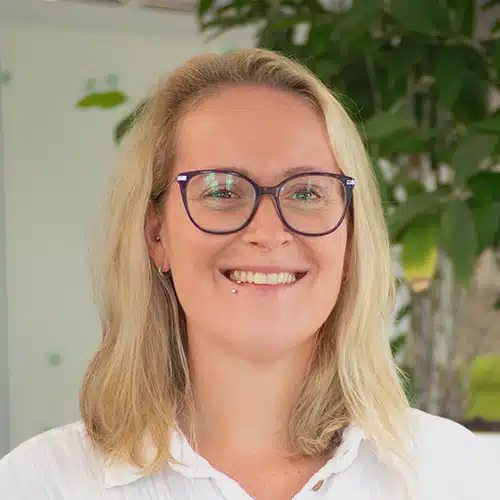 Portrait de Denise Raupp, Département Achats chez Debatin. Elle sourit gentiment, porte des lunettes et un piercing à la lèvre. Ses cheveux blonds tombent aux épaules, et elle porte un haut blanc devant un arrière-plan vert flou.