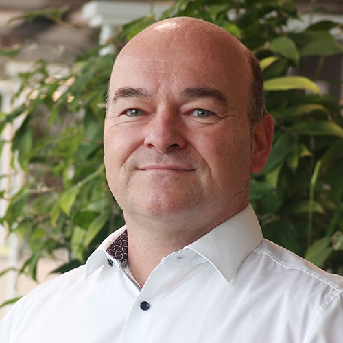 Portrait de Dr. Karsten Tauber. C’est un homme d’âge moyen avec la tête chauve et une chemise claire, souriant aimablement à la caméra. Une plante verte feuillue est visible en arrière-plan.