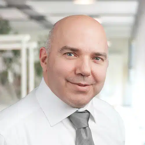 Portrait en buste de Helmut Roth, Département Achats chez Debatin. Il regarde directement la caméra avec un léger sourire. Homme chauve portant une chemise blanche et une cravate grise. L’arrière-plan flou suggère un environnement de bureau.