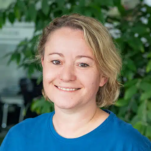 Gros plan de Michaela Breßer, travaillant comme Consultante commerciale / Service client chez Debatin. Une femme souriante avec des cheveux courts blonds portant un T-shirt bleu. Des plantes vertes floues sont visibles à l’arrière-plan.