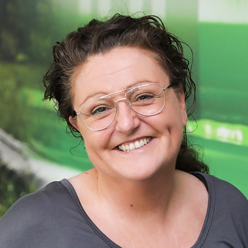 Cette image montre Nicole Klingler, Consultante technique des ventes chez Debatin. En gros plan, elle sourit, avec des cheveux bouclés, des lunettes et un haut gris.