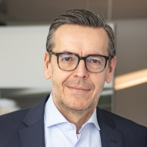 Portrait d’Oliver Kosalla, Responsable des achats chez Debatin. Il porte des lunettes modernes, une veste bleu foncé sur une chemise bleu clair, et regarde directement la caméra. Son visage montre une barbe soignée et des cheveux grisonnants.
