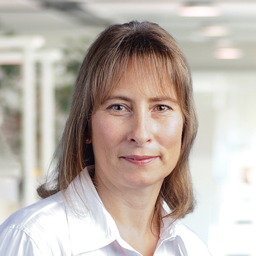 Vue détaillée de Sandra Däschner, Département Achats chez Debatin. Elle apparaît avec une expression ouverte et amicale et porte un chemisier blanc classique. Ses cheveux bruns mi-longs encadrent son visage devant un arrière-plan clair et flou.
