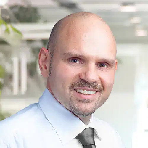 Portrait en buste de Thomas Harböck, Département Achats chez Debatin. Il sourit à la caméra, chauve avec une moustache soignée. Il porte une chemise claire et une cravate sombre, avec des accents verts flous à l’arrière-plan.