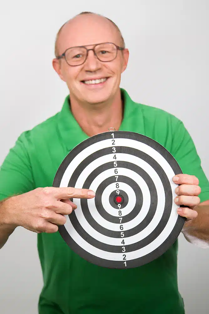 DEBATIN_Produktsparten_Headerbild_DEBADEINS_Gerald Frank_Zielscheibe Gerald Frank porte une chemise verte et tient une cible de fléchettes colorée.