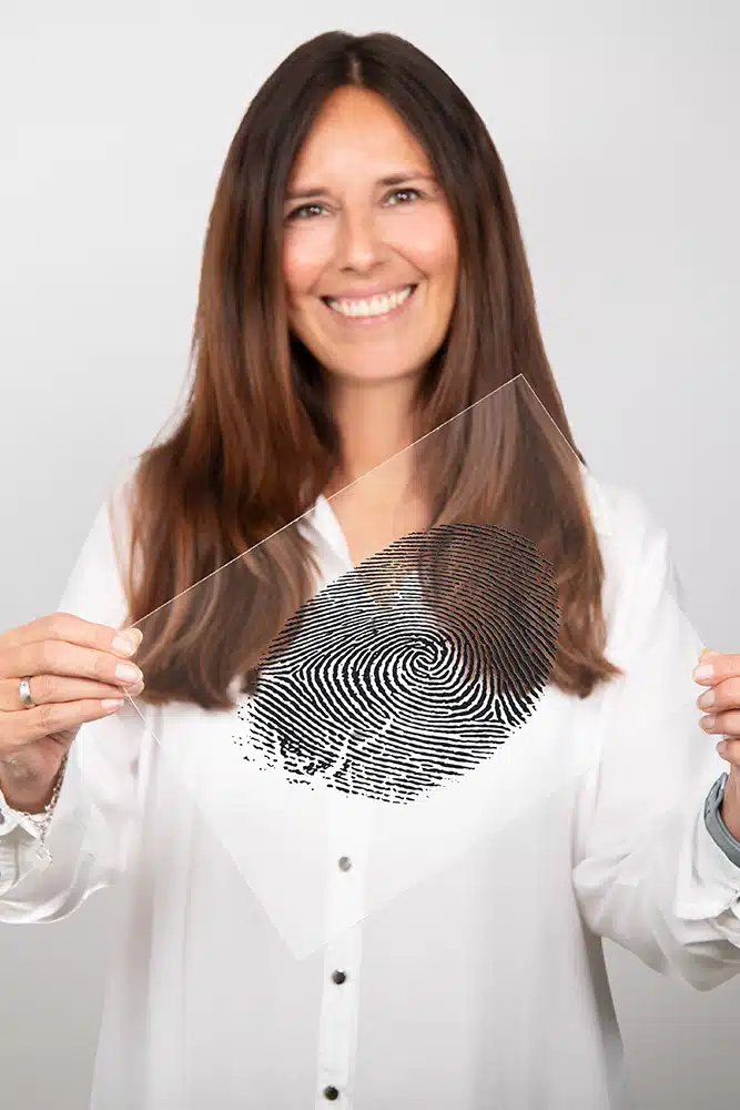 DEBATIN_Produktsparten_Headerbild_DEBADEINS_Hillenbrand_Fingerabdruck Gaby Hillenbrand porte un chemisier blanc et tient une empreinte digitale gravée dans une plaque en plexiglas.