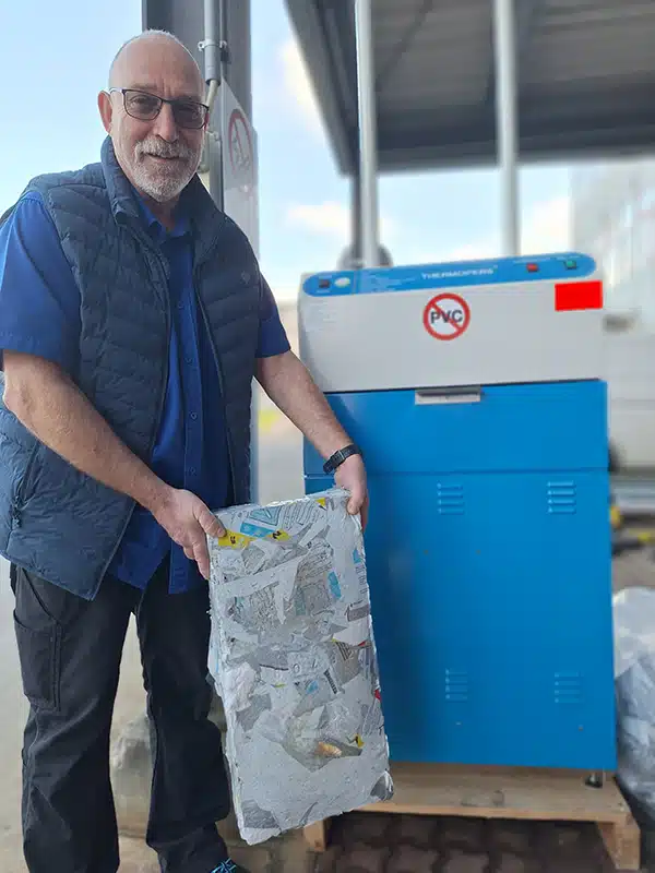 debatin_press_bioscientia Jürgen Heß, responsable de la gestion des matériaux chez Bioscientia Healthcare GmbH, se tient à côté de la presse de recyclage DEBATIN pour plastiques bleue. Il tient dans ses mains un bloc compacté de matériau plastique recyclé. À l’arrière-plan, on distingue une zone extérieure couverte d’un bâtiment industriel.