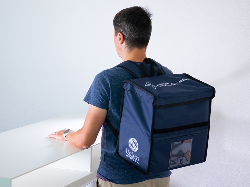 Une personne se tient devant une table blanche et porte un grand sac de transport bleu avec rabat, pochette transparente et logo blanc.