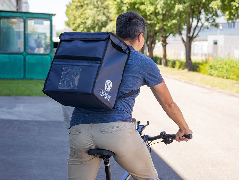 Une personne circule à vélo et porte un grand sac de transport bleu avec bordure noire, pochette transparente et logo blanc sur le dos.